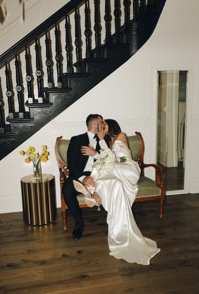 A couple in wedding attire kisses while sitting on a vintage loveseat in front of a staircase. A small table with yellow flowers is beside them.