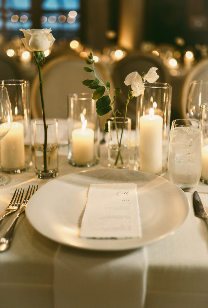 A table setting with a white plate, napkin, cutlery, and glasses. Candles in various heights and flowers in vases are arranged as centerpiece decor.