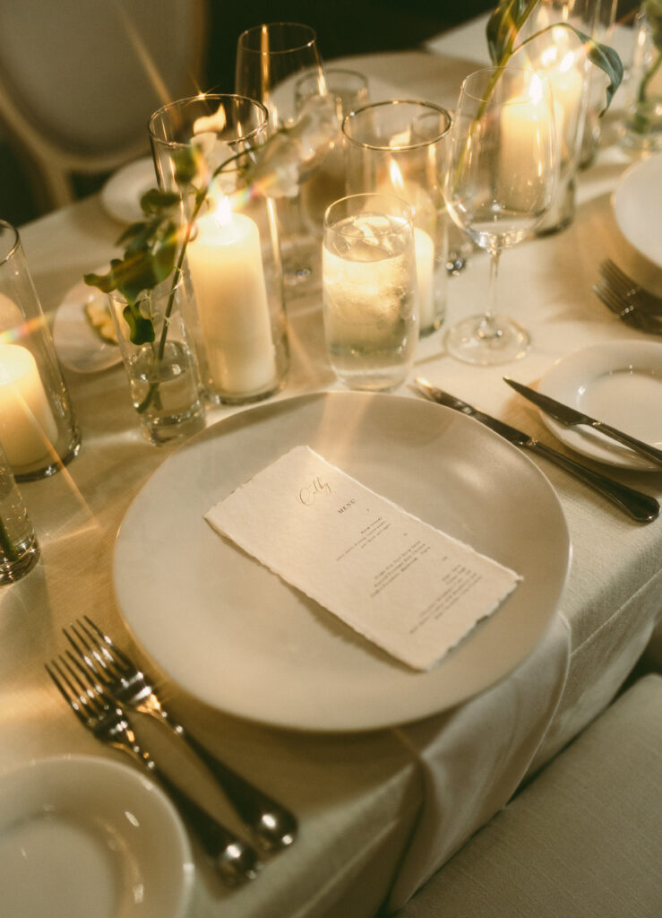 Elegant table setting featuring a menu on a plate, surrounded by candles, glasses, and cutlery on a cream tablecloth.