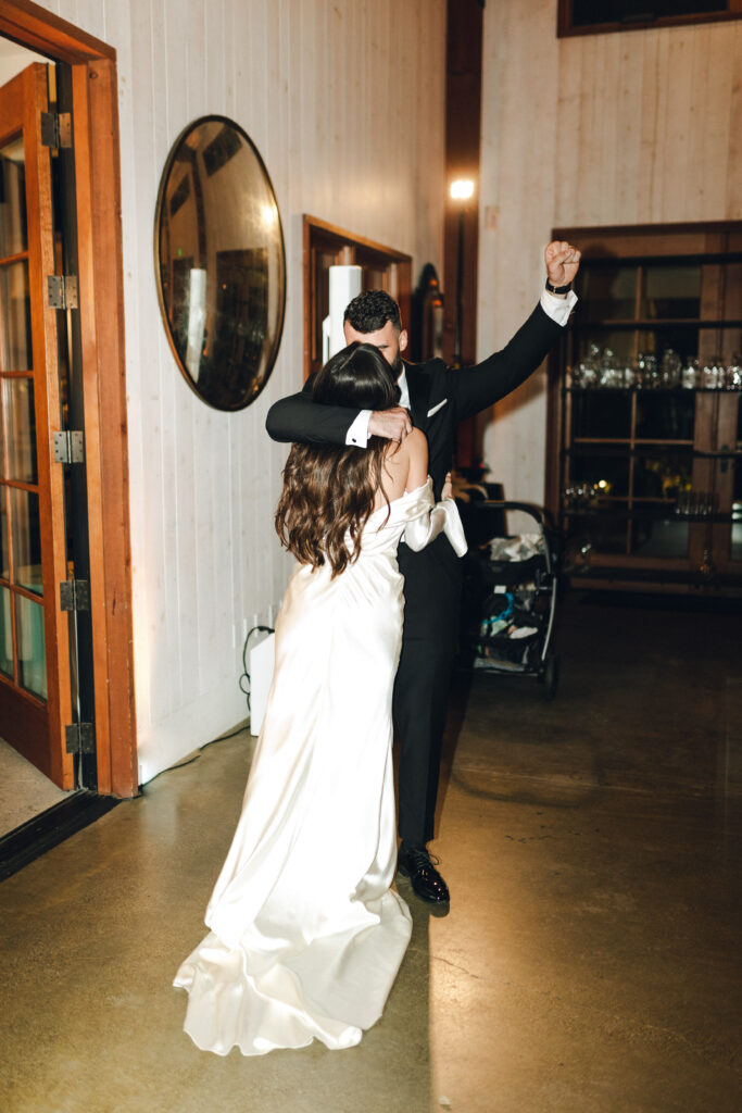 A couple dances indoors; the woman in a white dress and the man in a suit. The setting includes a round mirror and glassware on shelves.