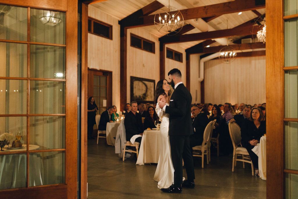 A bride and groom share a dance in a rustic reception hall, surrounded by seated guests.