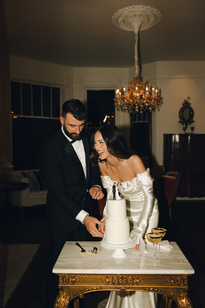 A couple in formal attire are smiling and cutting a white cake on a small table with two glasses nearby, in a dimly lit room with a chandelier.