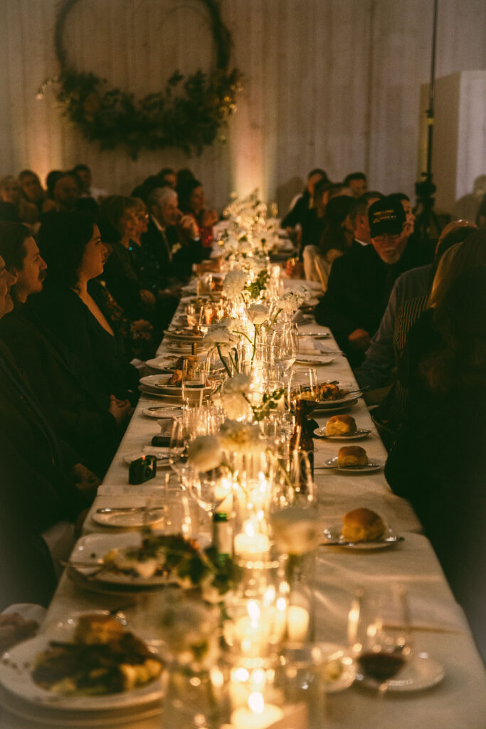 A long table set for a dinner event, with plates, glasses, and candles. People are seated on both sides, and a decorative wreath hangs on the wall in the background.