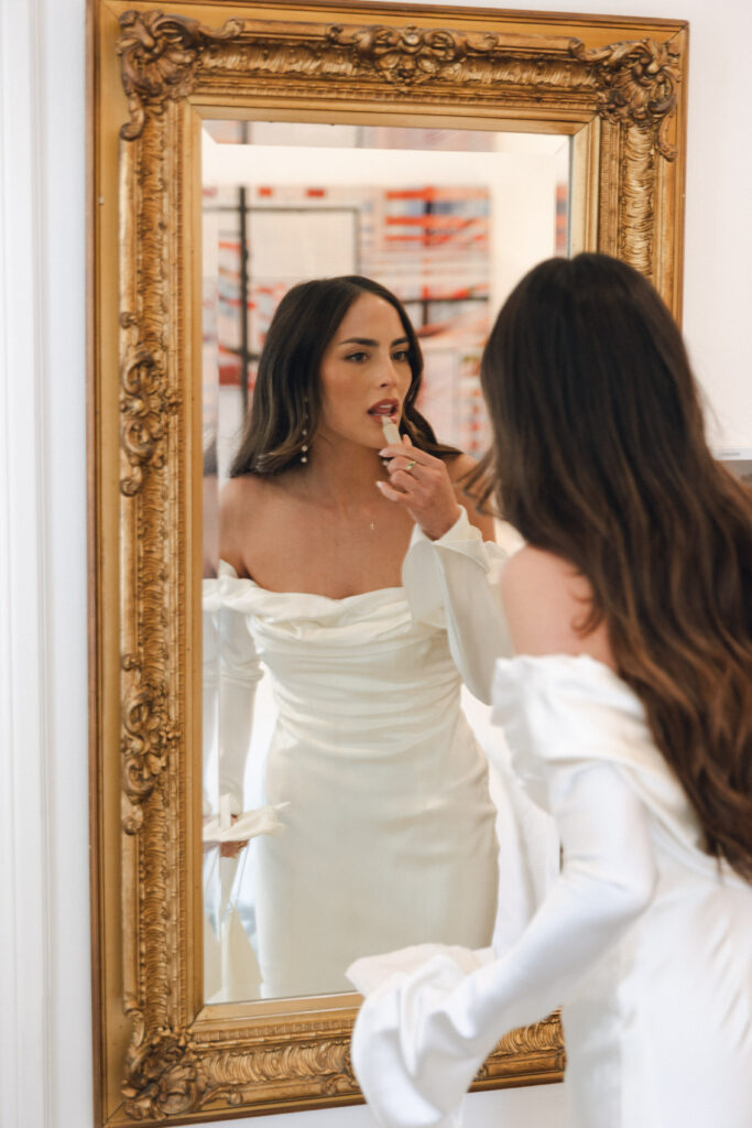 A woman in a white off-shoulder dress applies lipstick while looking in a large ornate mirror.