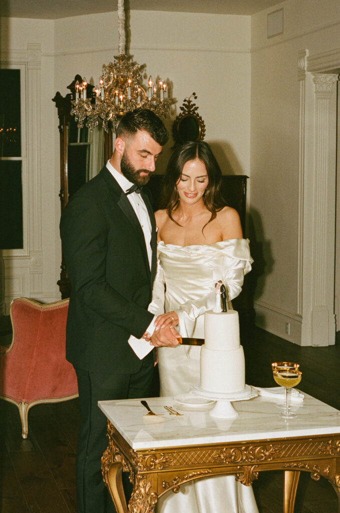 A couple in formal attire cuts a white cake on a marble table in a room with a chandelier and antique decor.