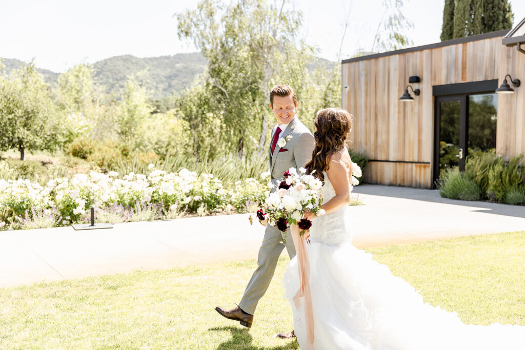 A bride and groom walk outdoors on grass near a wooden building and garden. The bride holds a bouquet of white and maroon flowers. It's a sunny day.