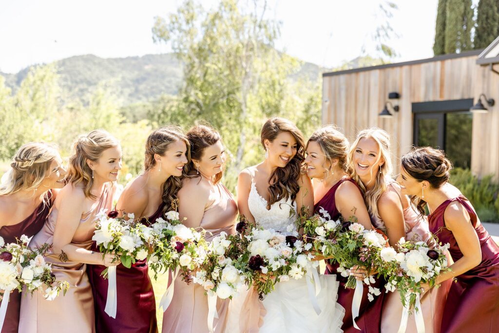 Bride surrounded by bridesmaids holding bouquets, posing outdoors in front of a scenic backdrop.