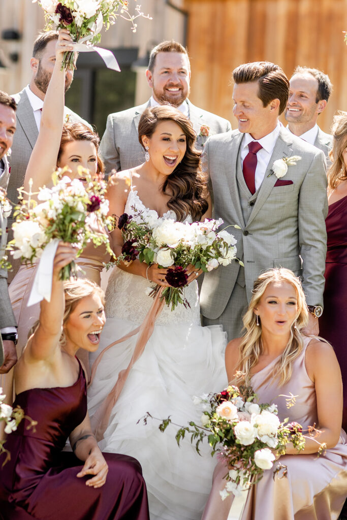 A wedding party poses joyfully; the bride and groom are surrounded by bridesmaids and groomsmen, all smiling and holding bouquets.