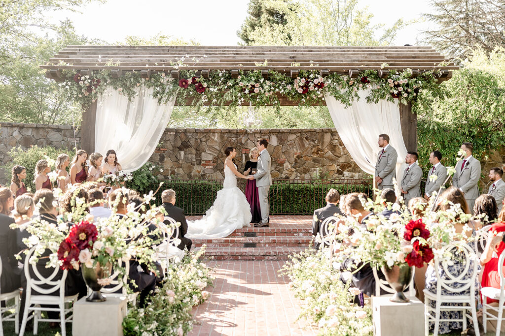 A bride and groom stand under a decorated wooden pergola during an outdoor wedding ceremony, surrounded by the wedding party and seated guests.