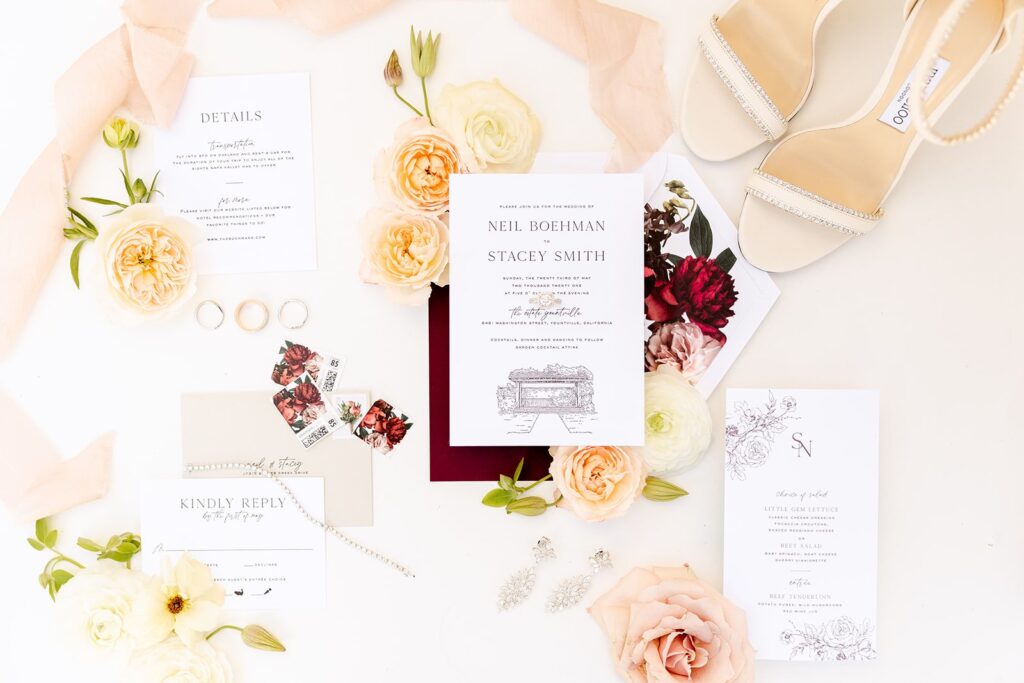 Flat lay of wedding invitation suite with floral decorations, rings, reply cards, and shoes on a white background.