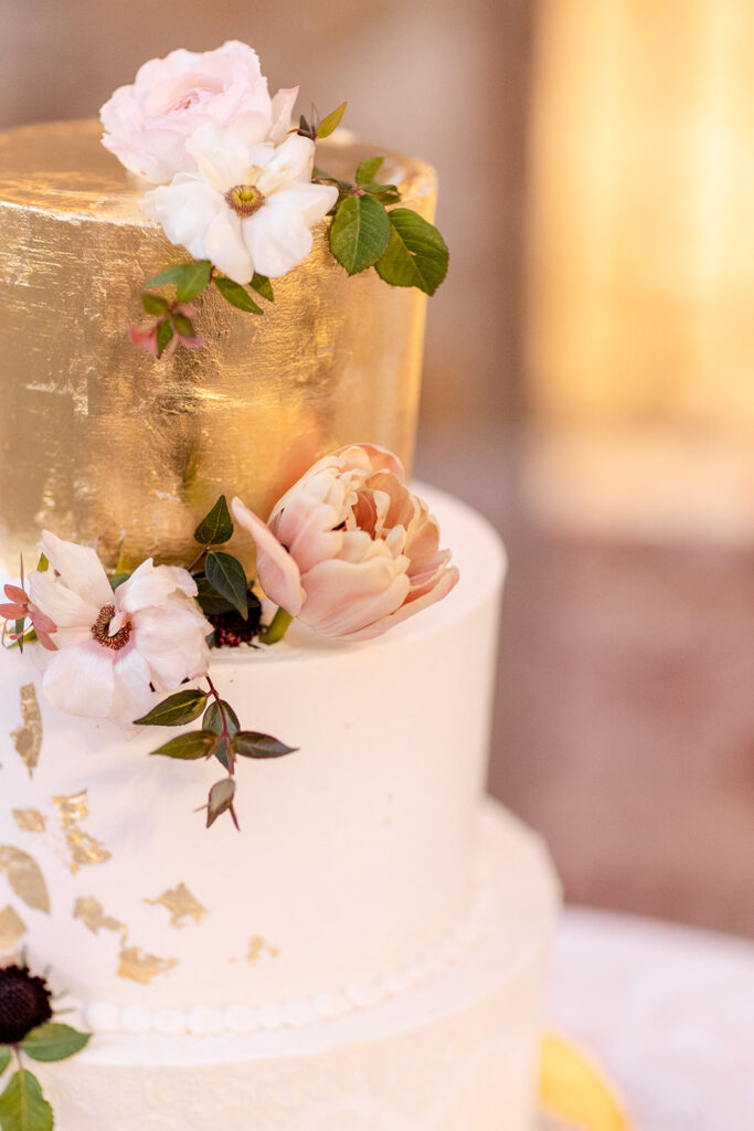 A two-tiered wedding cake with a golden top tier, adorned with white and blush flowers and green leaves.