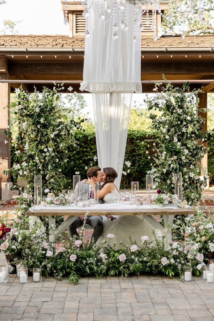 A couple in wedding attire kiss while seated at a decorated outdoor table, surrounded by greenery and flowers.