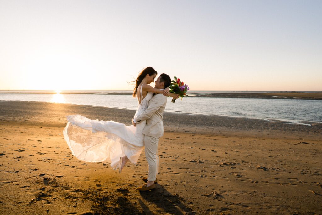 A groom in a light suit lifts a bride carrying a bouquet on a sandy beach at sunset. Her dress flows in the breeze.