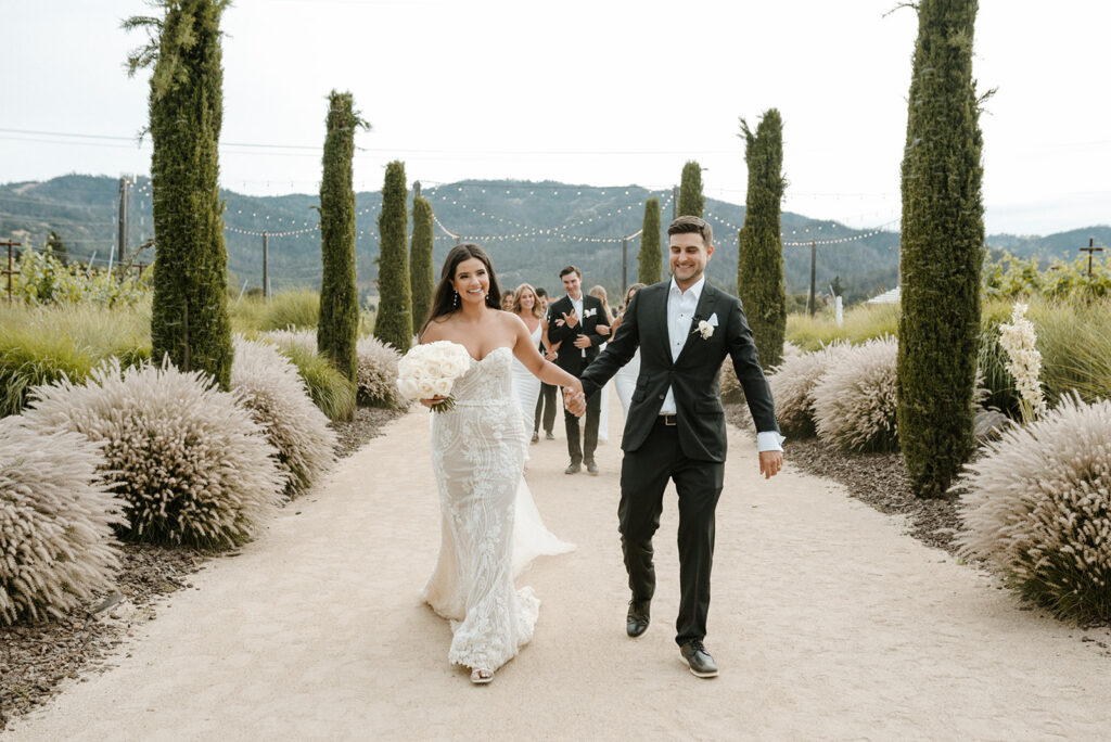Bride and groom walk joyfully down a garden path, followed by their wedding party, surrounded by tall greenery and mountains in the background.