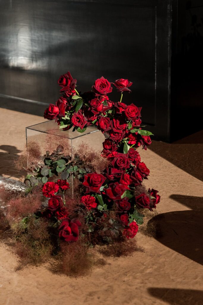 Elegant arrangement of red roses in a transparent square vase, set against a dark background with dappled shadows on a beige surface.