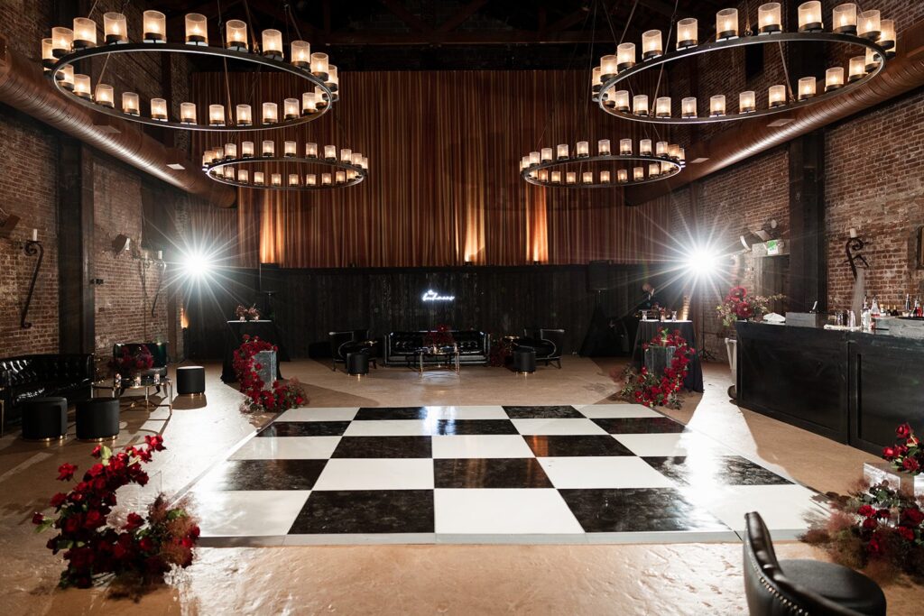 Dimly lit ballroom with black and white checkered dance floor, surrounded by brick walls, floral arrangements, and large circular chandeliers overhead.