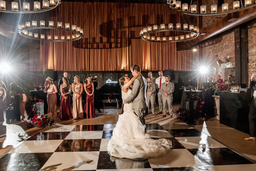 Bride and groom dance on a checkered floor at a wedding reception, surrounded by guests and lit by large circular chandeliers.