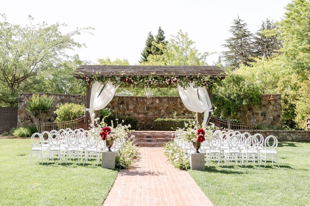 Outdoor wedding ceremony setup with white chairs, floral arrangements, and a wooden pergola draped with sheer fabric and flowers, on a grassy lawn with trees in the background.