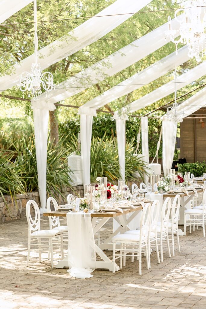 Outdoor wedding reception setup with white chairs, a long decorated table, floral centerpieces, and hanging chandeliers under a clear tent with greenery in the background.