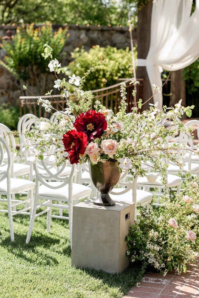Flower arrangement with red and white blooms in a vase atop a pedestal, surrounded by greenery. Rows of white chairs are in the background, set outdoors with a draped structure.