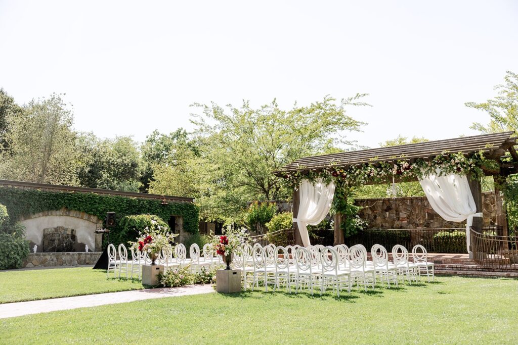 Outdoor wedding setup with white chairs arranged in rows facing a wooden pergola adorned with white drapes and flowers. Greenery surrounds the scene on a sunny day.