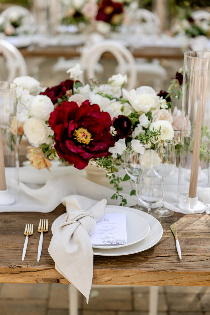 Elegant table setting with white plate, beige napkin, menu, gold flatware, and abundant floral centerpiece of red and white blooms, on a rustic wooden table.