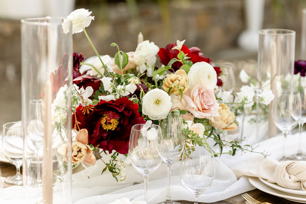 Elegant floral centerpiece with red, white, and pink flowers surrounded by clear wine glasses and candles on a white tablecloth.