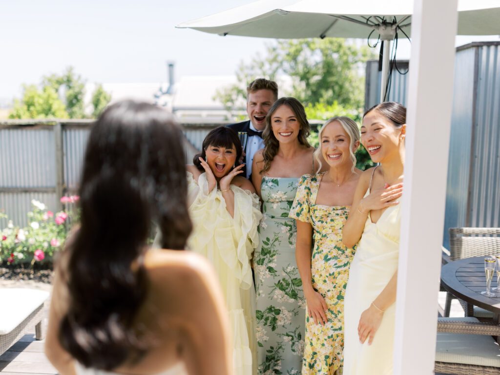 A person with long hair is in the foreground, while a group of five smiling people in colorful outfits stands under an outdoor umbrella looking towards them.