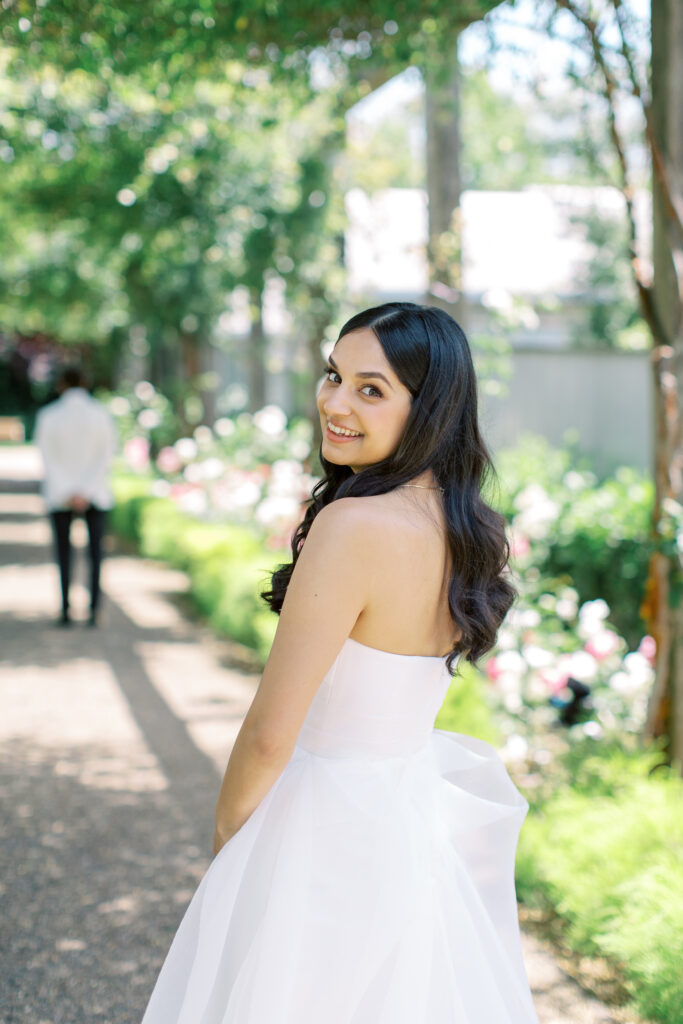 A woman in a strapless white dress smiles, standing on a garden path with sunlight filtering through trees.