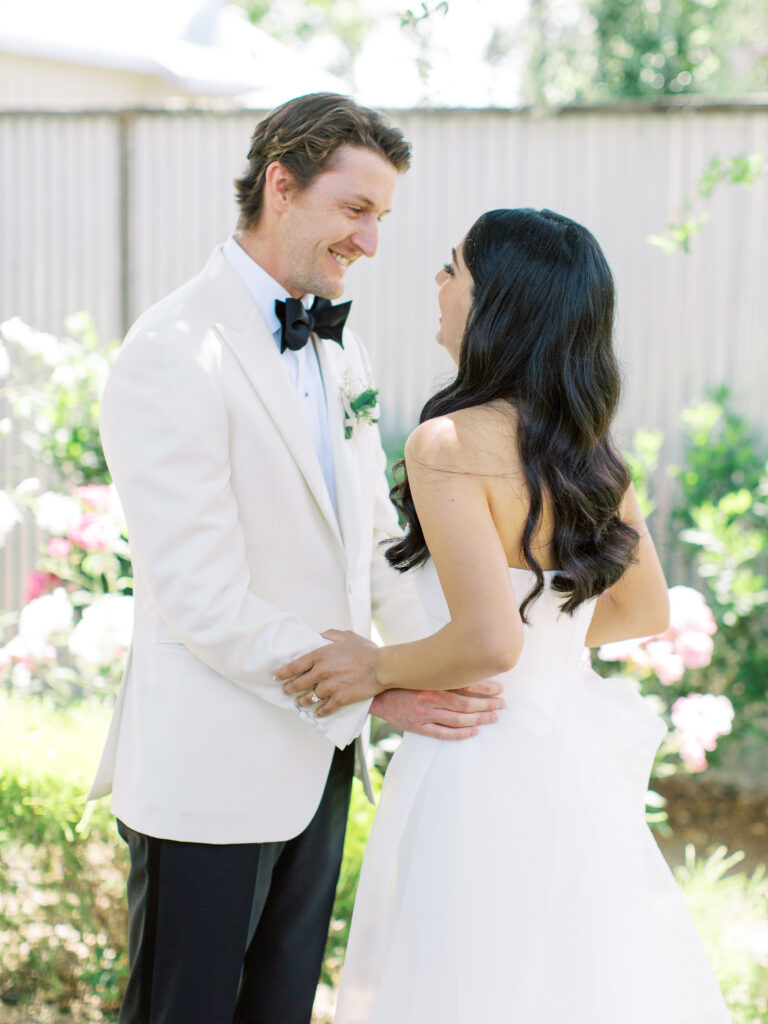 A bride and groom smile at each other in a garden setting, both wearing formal attire.