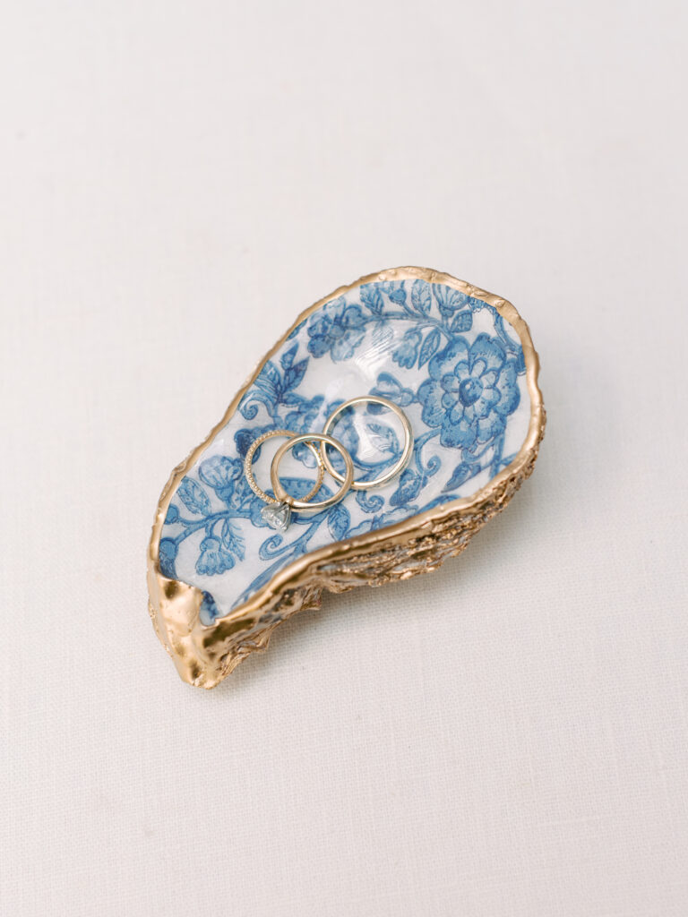 Two rings rest in a decorative oyster shell dish with blue floral patterns and gold edges on a light surface.