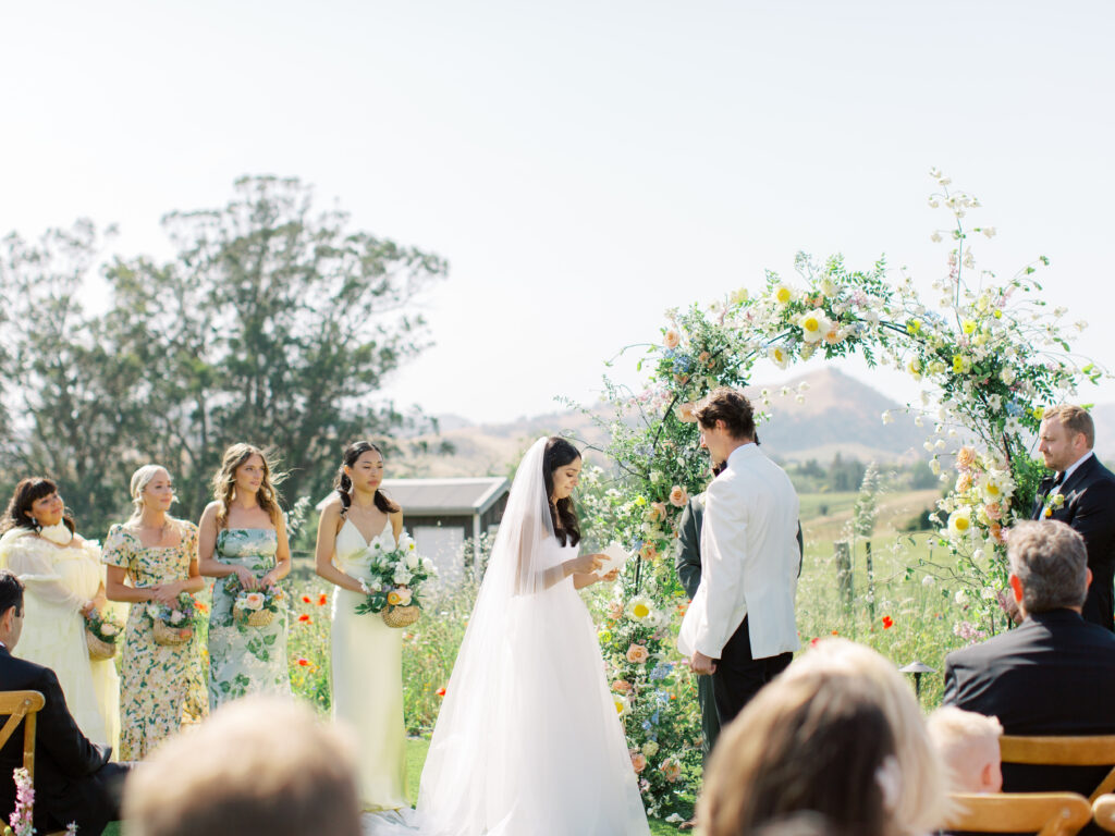 Bride and groom stand under a floral arch during an outdoor wedding ceremony, with bridesmaids in floral dresses nearby and guests seated in the foreground.