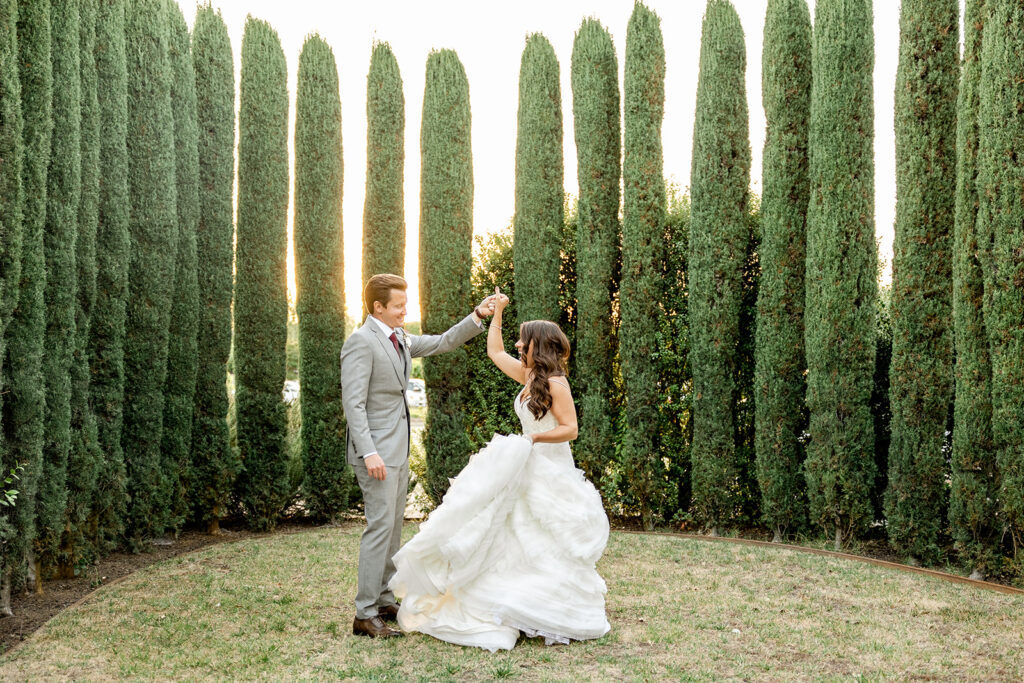 The couple dances on grass, surrounded by tall, narrow trees. The woman wears a white gown, and the man is in a gray suit. Sunlight filters through the trees in the background.