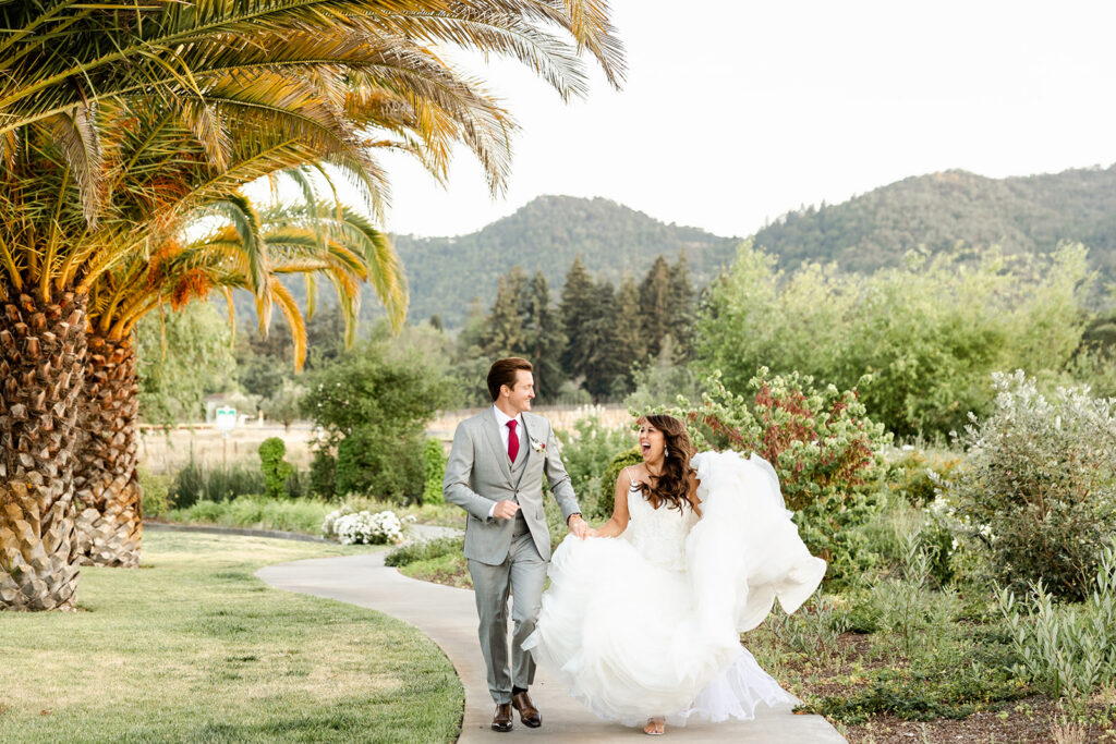 A bride in a white dress and groom in a gray suit walk joyfully along a garden path, with mountains in the background.