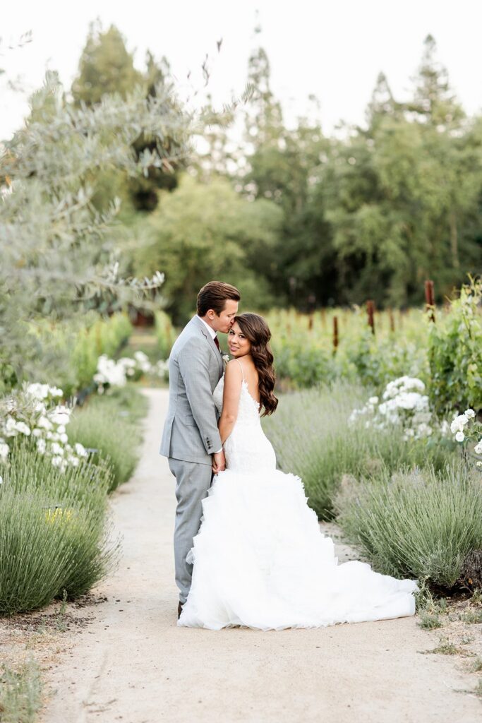 A couple stands on a path in a garden, with the woman in a white wedding dress and the man in a gray suit, surrounded by greenery and white flowers.