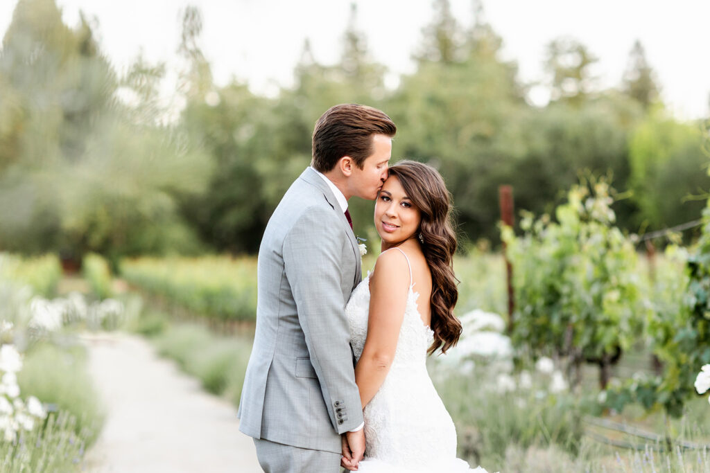 A couple embraces on a path in a garden. The man is wearing a gray suit, while the woman is in a white dress. They are surrounded by greenery and flowers.