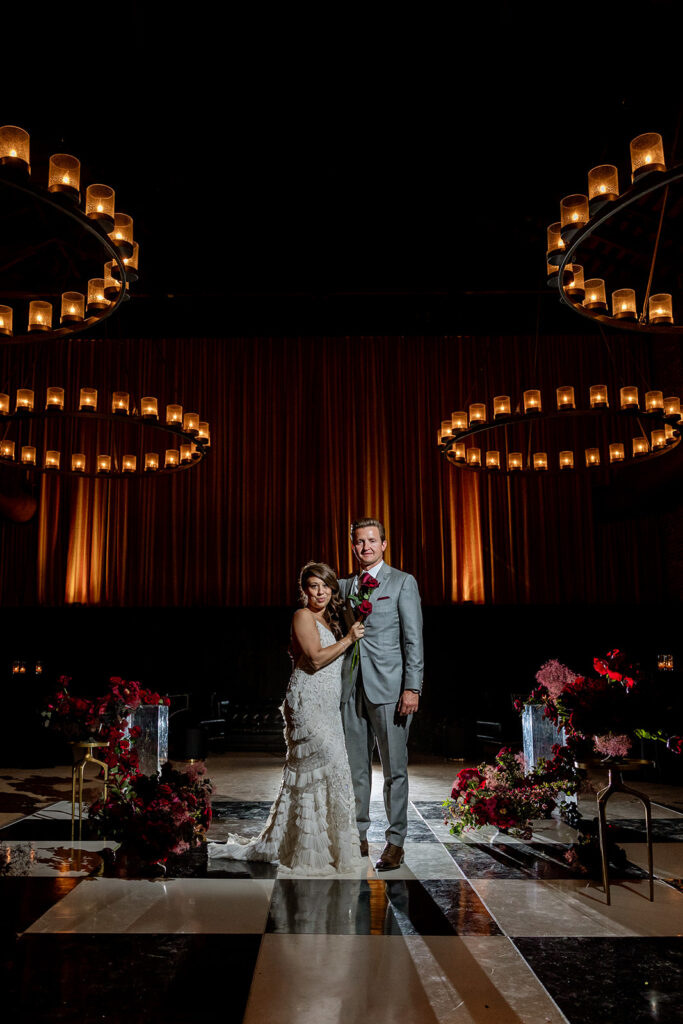 A bride and groom stand together on a dark, checkered floor with floral arrangements, under circular chandeliers with lit candles.