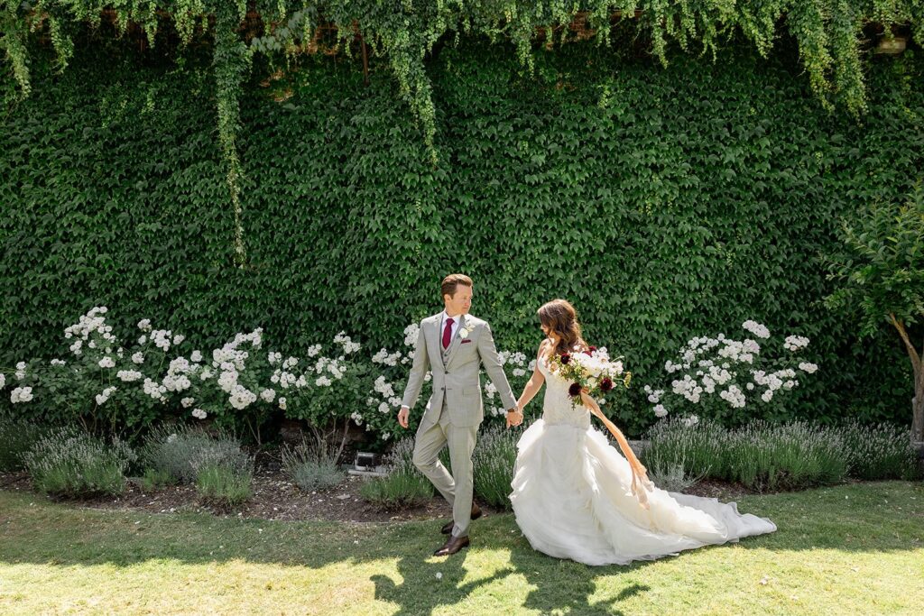 Bride and groom walking hand in hand in front of a lush green ivy wall and white flowers under bright sunlight.