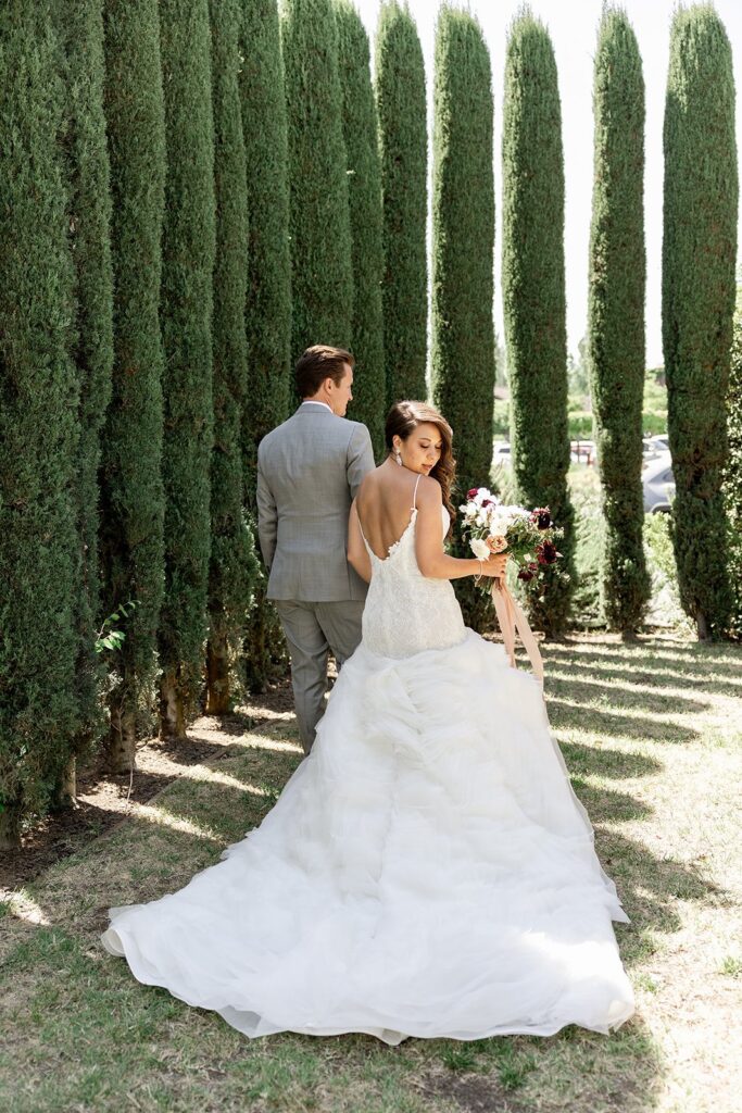 Bride and groom walking between tall trees, she in a white gown holding a bouquet, he in a gray suit.