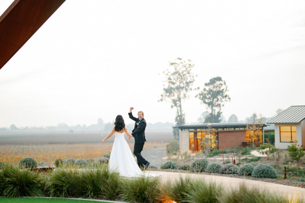 Bride and groom joyfully dance on a path near landscaped bushes, with a modern building and open field in the background.