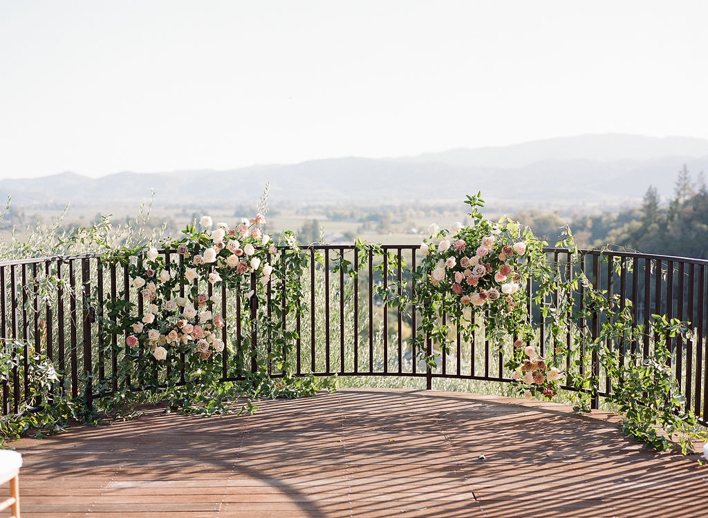Outdoor balcony with a metal railing decorated with pink and white flowers and greenery, overlooking a scenic landscape with hills in the background.