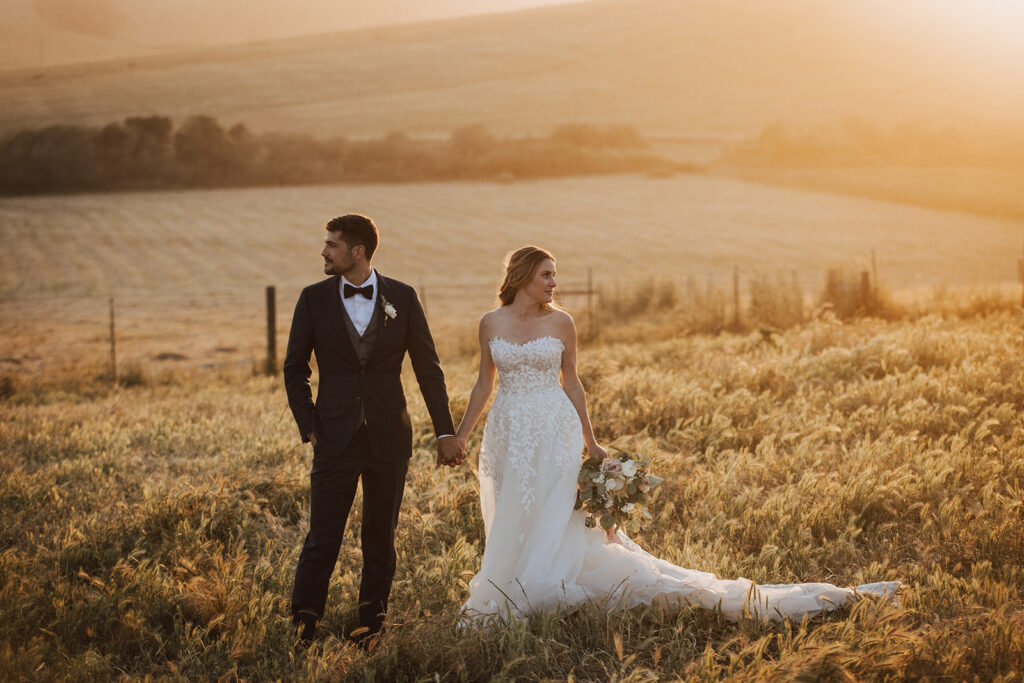 A bride and groom stand in a sunlit field, holding hands. The groom wears a dark suit and bow tie, while the bride wears a white gown and holds a bouquet.