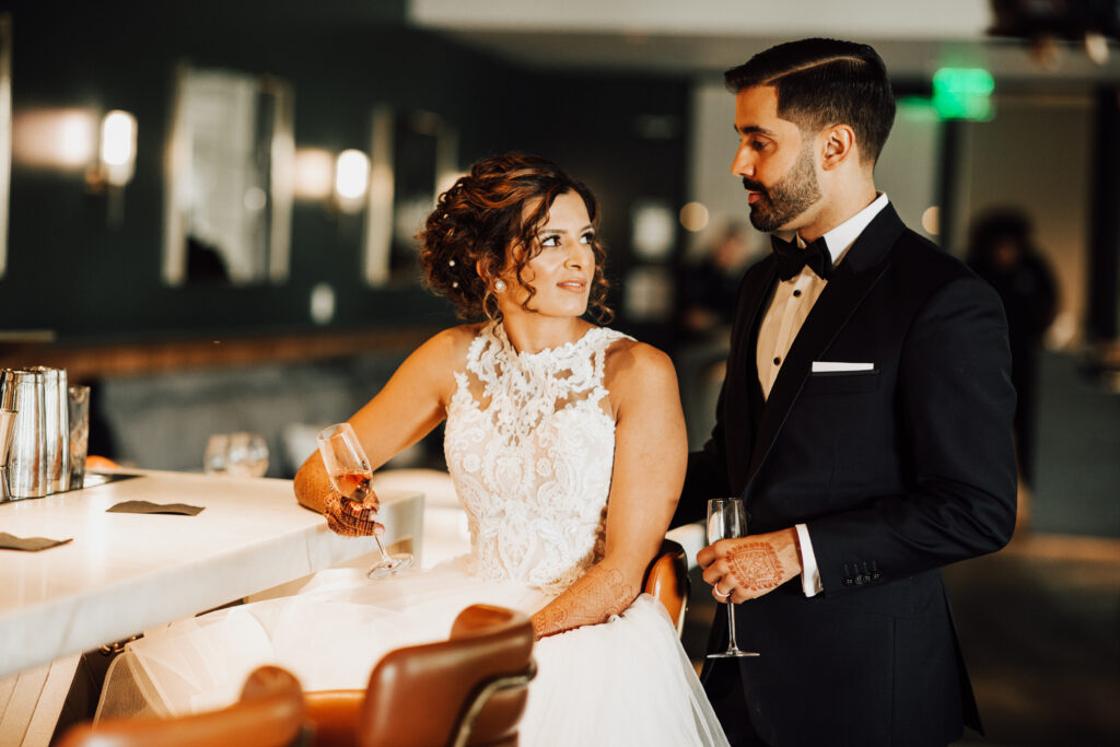 Bride and groom in formal attire stand by a bar, holding champagne glasses.