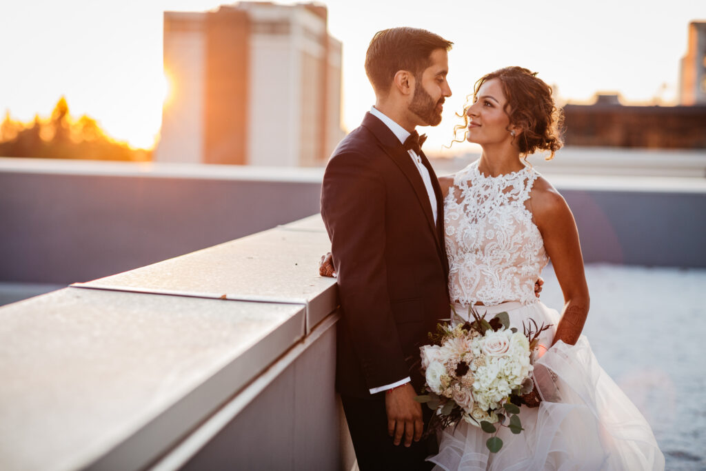 A bride and groom stand close and smile at each other on a rooftop at sunset, with the groom in a suit and the bride holding a bouquet.