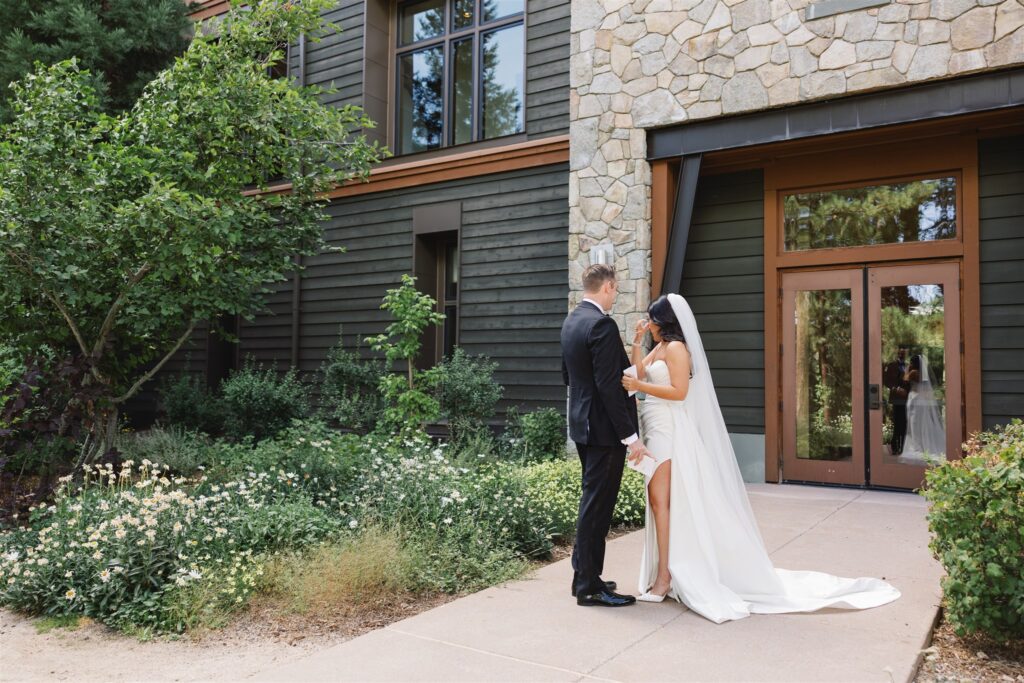 A bride and groom stand together in front of a building with stone and wood siding, surrounded by greenery and flowers.