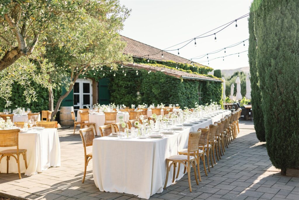 Outdoor event space with long tables covered in white tablecloths, adorned with white floral arrangements and surrounded by wooden chairs, set under string lights, next to a vine-covered building.