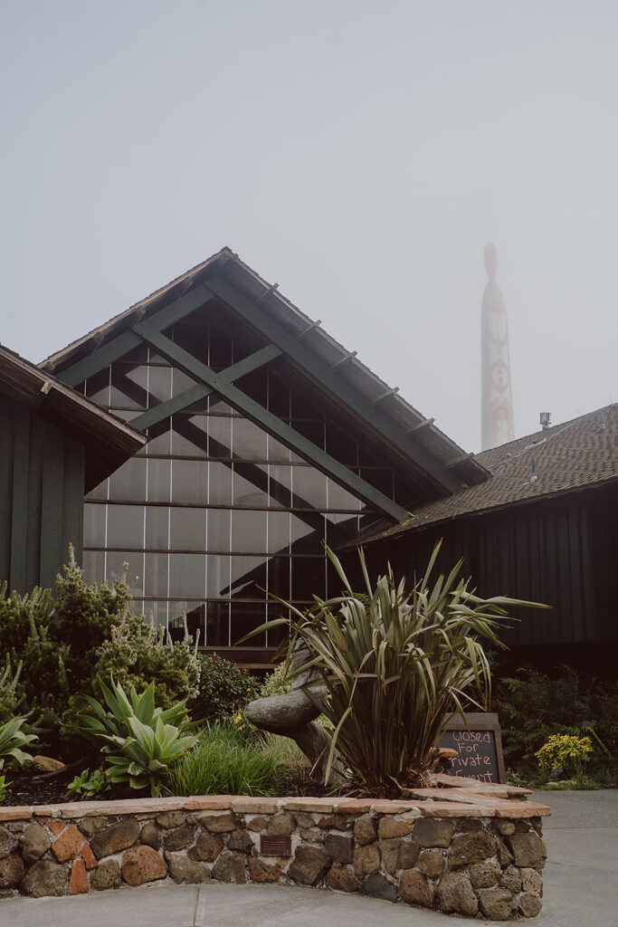 A-frame building with large glass windows, surrounded by lush plants. A chalkboard sign reads "Closed for private party" near a stone border. A tall wood structure is visible in the background.