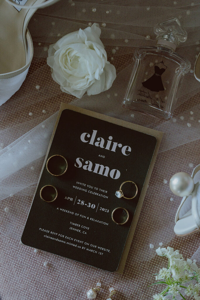 Wedding invitation with rings and perfume bottle on a textured surface, surrounded by white flowers and pearls.