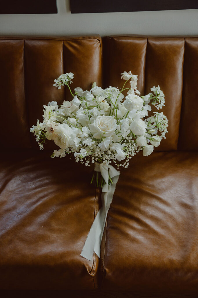 A bouquet of white flowers, including roses and baby's breath, tied with a white ribbon, placed on a brown leather couch.