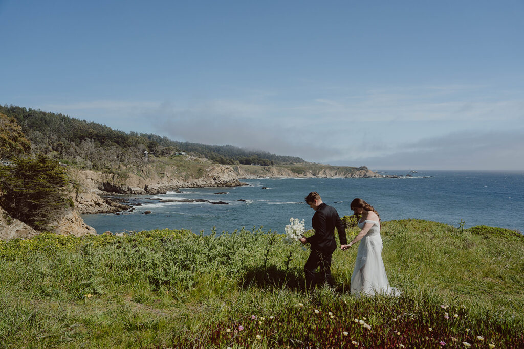 Bride and groom holding hands walking on a grassy hill by the ocean, with cliffs and a clear blue sky in the background.